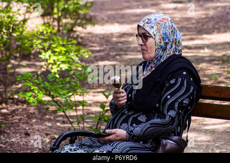 Niska Banja, Serbien - Juli 02, 2018: Muslimische alte Frau auf der Bank entspannen und isst Eis cram im Schatten im Naturpark. Urlaub und Genuss des Sommers Stockfoto