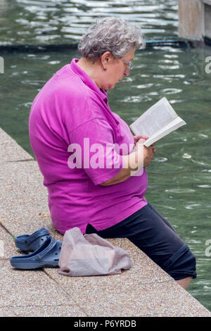 Niska Banja, Serbien - Juli 02, 2018: Alte Frau mit Buch mit Beinen in gesundes Wasser im Kurpark. Urlaub und Genuss des Sommers Konzept Stockfoto