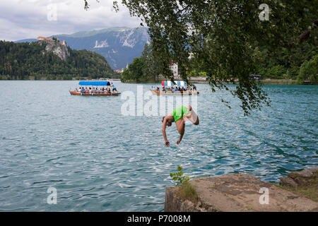 Ein Junge Tauchgänge in das kühle Wasser des Sees Bled mit Pletna Boote, die Touristen über den See, am 18. Juni 2018 in Bled, Slowenien. Stockfoto