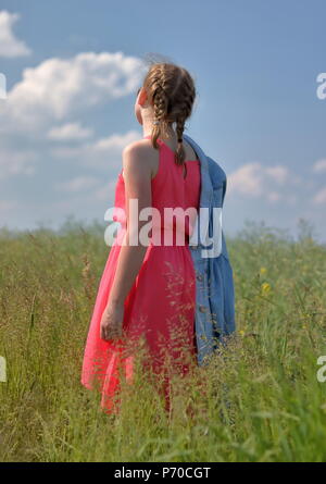 Eine weiße Haut junge blonde Haare Mädchen steht wieder auf Wiese in rosa Sommer sleevless Kleid, sonnigem Wetter, blauer Himmel mit weißen Wolken Stockfoto