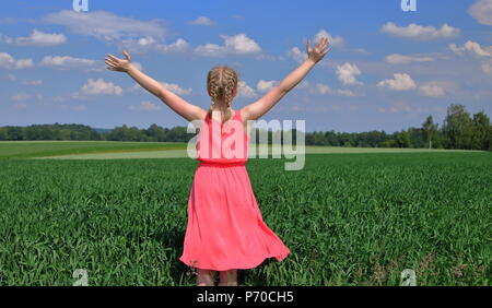 Junge Frau, blonde Haare, kurze Zöpfe, steht auf dem Rücken im grünen Feld, auf dem Land, Sommer, im rosa Kleid mit offenen Rams gekleidet, Freuden Stockfoto