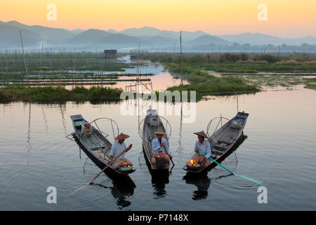 Drei Fischer warm halten in ihren langen Schwanz Fischerboote in der Dämmerung in der Nähe der schwimmenden Gärten am Inle See, Shan Staat, Myanmar (Birma), Asien Stockfoto