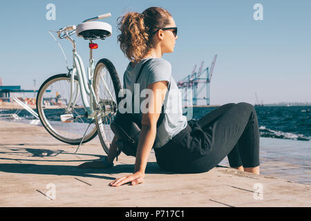 Junge Frau sitzt auf Holz- Damm auf See in der Nähe ihrer Stadt Fahrrad suchen. Frauen tragen, Rucksack und blaues t-shirt Ausruhen nach Radtour rund um Stockfoto