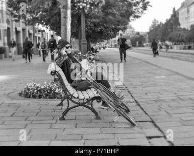 KOSICE, SLOWAKEI - Oktober 02, 2017: Schöne junge Frau sitzt mit Smartphone und Fahrrad auf der Bank in der Altstadt in Schwarz und Weiß. Kosice Stadt w Stockfoto