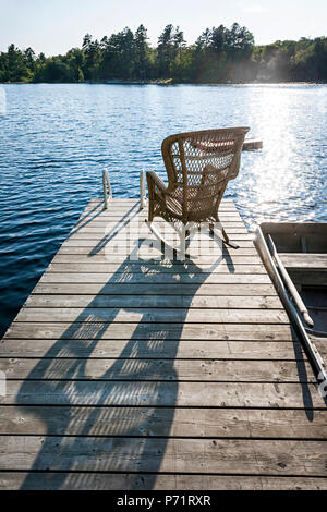 Rattan Schaukelstuhl auf Holzterrasse im Sommer am kleinen See werfen lange Schatten Stockfoto