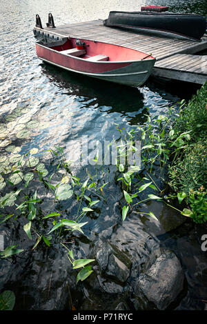 Red Boat an der hölzernen Dock auf See in Cottage Country mit Vordergrund der Felsen und Pflanzen Stockfoto