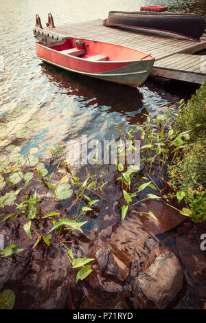 Red Boat an der hölzernen Dock auf See in Cottage Country mit Vordergrund der Felsen und Pflanzen Stockfoto