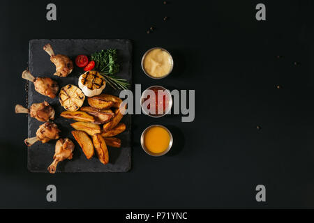 Blick von oben auf die leckere gebackene Kartoffeln mit gegrillter Knoblauch und Huhn, Kräuter und Saucen auf Schwarz Stockfoto