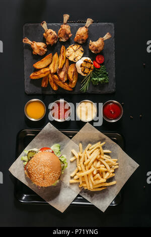 Blick von oben auf die Pommes Frites mit leckeren Burger auf Fach, verschiedene Saucen und Schiefer mit gerösteten Kartoffeln, gegrilltem Gemüse und Chicken Wings auf Schwarz Stockfoto