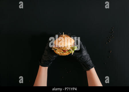 7/8 shot der Hände in Handschuhe Holding leckere Burger mit der Türkei, Gemüse und Caesar Dressing auf Schwarz Stockfoto