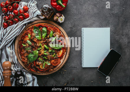 Blick von oben auf die frisch gebackene Pizza mit Notebook und Smartphone auf konkrete Tabelle Stockfoto