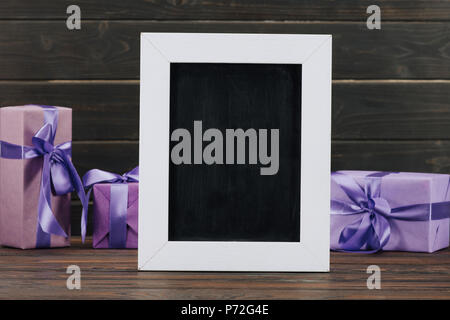 Leere Schiefertafel in Rahmen mit Geschenkboxen gegen die Holzwand Stockfoto