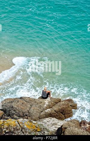 Im mittleren Alter blonde Frau, sitzen auf den Felsen durch einen Cornish blue sea Porthcurno Cornwall England UK umgeben Stockfoto