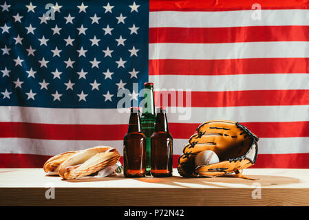 Nahaufnahme der Bierflaschen, Hot Dogs, Leder Handschuh und baseball ball auf Holztisch mit us Flag hinter Stockfoto