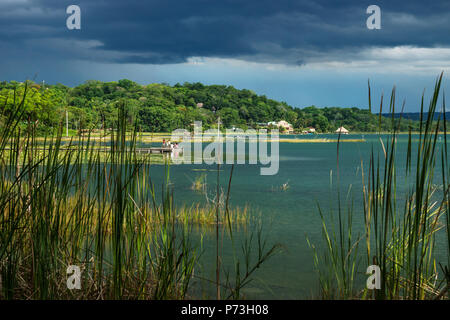 Blick auf das Dorf mit Dock am Seeufer entlang mit dunklen Blau cloudscape und Sonnenschein, El Remate, Peten, Guatemala Stockfoto