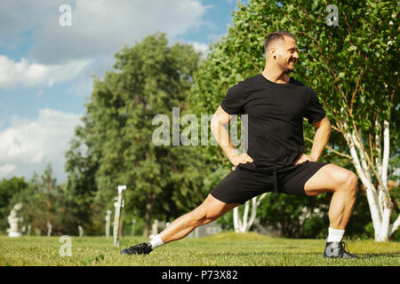 Junger attraktiver Mann in Schwarz Sportbekleidung, ausfallschritt Outdoor im Park. Stockfoto