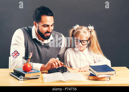 Intelligente männliche Lehrer in graue Weste erklären, neues Material auf der primären Schüler Mädchen am Tisch mit Büchern Stockfoto