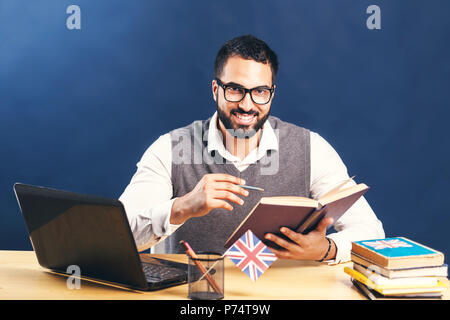 Dunkelhaarige bärtiger Mann Englisch lernen, tragen graue Strickjacke Weste und unberührten weißen Shirt, hart Arbeiten am Schreibtisch mit Laptop vor der schwarzen Wand Stockfoto