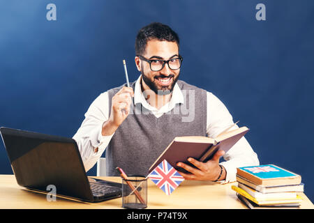 Dunkelhaarige mann Englisch lernen, tragen graue Strickjacke Weste und unberührten weißen Shirt, lächelnd im Büro Schreibtisch mit Laptop vor der schwarzen Wand Stockfoto