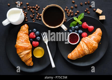 Frühstück mit Croissants, Honig, Marmelade, Kaffee schwarz, creme und frische Beeren auf schwarzem Hintergrund. Ansicht von oben, flach Komposition legen Stockfoto