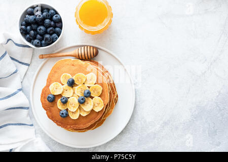 Buttermilch Pfannkuchen mit Banane, Blaubeeren und Honig auf konkreten Hintergrund. Ansicht von oben und kopieren Sie Platz für Text. Blick von oben auf die Pfannkuchen mit Beeren auf Stockfoto