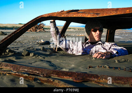 Mann sitzt auf verrosteten Autowrack vergraben im Sand am Strand Stockfoto