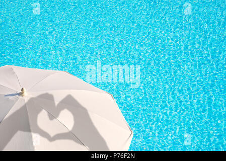 Schatten der Hände bilden ein Herz auf einem weißen Sonnenschirm, blau Schwimmbad Wasser Hintergrund, Sommer Konzept Stockfoto