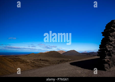 Schotterstraße in die große Leere und Einsamkeit der Lanzarote schwarz gefrorener Lava vulkanische Wüste und einige schlafende Vulkane im Hintergrund. Steinmauer in der rechten Seite des Bildes Stockfoto