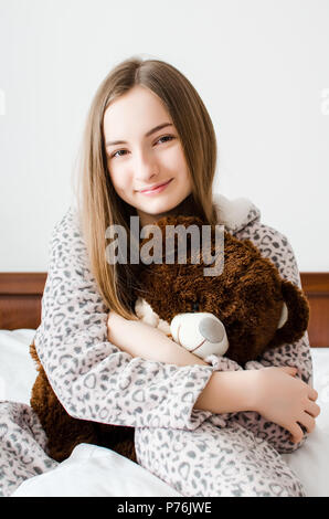 Glückliche junge Frau mit blondem Haar sitzen auf dem Bett in ihrem Schlafzimmer in einer Umarmung mit einem stofftier Spielzeug. Favorite brauner Teddybär in der Hand o Stockfoto