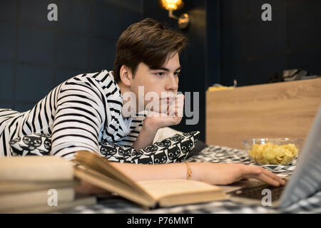 Attraktive junge männliche liegend auf Bett mit Bücher und Schüssel von Chips und mit Laptop Computer. Stockfoto