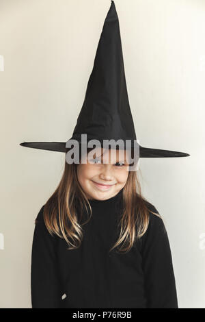 Lächelnd kleine blonde Mädchen mit den schwarzen Hexe Kostüm über weiße Wand, close-up Studio Portrait Stockfoto