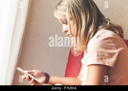 Schockiert Europäische blonde teenage Mädchen in rosa t-shirt mit einem Smartphone, einem Indoor closeup Profil Portrait Stockfoto