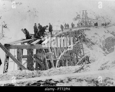 . Englisch: Crew arbeiten auf Holz gestellbrücke Verbindungstunnel Berg mit dem Südportal des Tunnels beim Bau der White Pass & Yukon Railroad, Alaska, Ca. 1899. Englisch: Legende auf Bild: "Brücke an Tunnel White Pass und Yukon Route" Bild im Hegg Album 2, Seite 26. Themen (LCTGM): Berge - Alaska; Eisenbahntunnel - Alaska - Tunnel Mountain; Railroad bridges - Alaska - Tunnel Mountain; Gerüste - Alaska - Tunnel Mountain; Eisenbahn Bau & Wartung - Alaska - Tunnel Mountain Themen (LCSH): Tunnel Mountain (Alaska); WHITE PASS AND YUKON ROUTE (Hart). Stockfoto