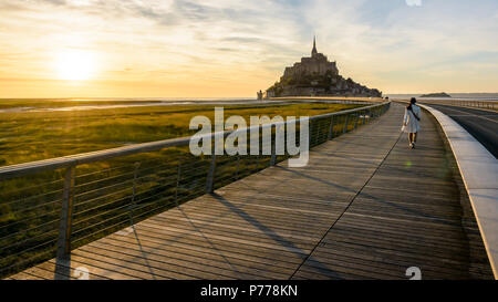 Panoramablick auf den Mont Saint-Michel tidal Island in der Normandie, Frankreich, bei Sonnenuntergang mit einer Frau zu Fuß in die Stadt auf dem hölzernen Steg. Stockfoto