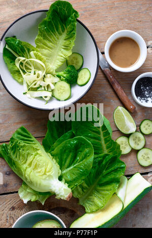 Zutaten zum Kochen grüner Salat auf hölzernen Tisch. Blätter Römersalat, Gurke, Avocado, Zucchini, Sesam und Soße auf einem Holztisch, Ansicht von oben Stockfoto