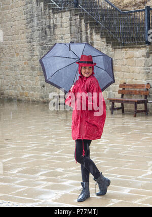 Junge glücklich attraktive hübsche Frau im Regen stehen mit einem Regenschirm in roter Kleidung. Stockfoto