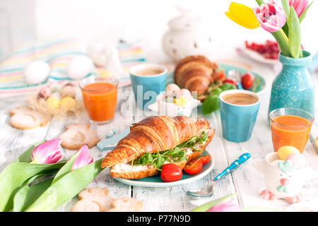 Hintergrund mit verschiedenen Farben. Eine Familie Frühstück mit Croissants mit Rucola und Käse und aromatischen Kaffee, Eier in verschiedenen Farben, helle Gerichte und Ostern Dekoration, Keramik Kaninchen Stockfoto