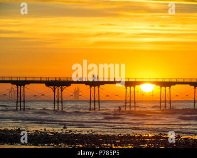 Redcar Offshore-Windfarm über einen Abschnitt der Salburn Pier während eines Sommer Sonnenuntergang gesehen, mit Menschen zu Fuß auf dem Pier und ein Surfer reiten sein Board Stockfoto