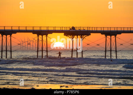 Redcar Offshore-Windfarm über einen Abschnitt der Salburn Pier während eines Sommer Sonnenuntergang gesehen, mit ein paar sitzt auf dem Pier und ein Surfer unter Reiten Stockfoto