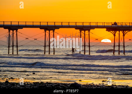 Redcar Offshore-Windfarm über einen Abschnitt der Salburn Pier während eines Sommer Sonnenuntergang gesehen, mit ein paar sitzt auf dem Pier und ein Surfer unter Reiten Stockfoto