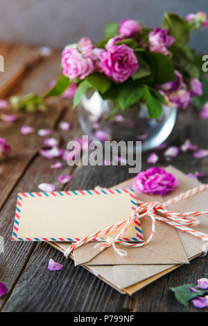 Vintage Konzept - Bouquet von Welke tee Rosen in Vase, Stapel alter Briefe in Umschläge und leere Grußkarte auf dem alten Holz- rustikalen Hintergrund. Stockfoto