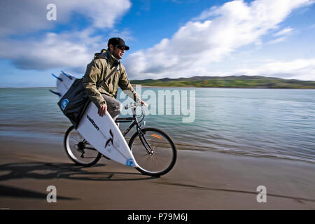 Surfer Reiten Fahrrad am Sandstrand zu Wellen, während Surfbrett in Raglan, Neuseeland Stockfoto