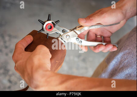 Die Bohrungen mit Locher in Leder Werkstatt. Männliche Hände mit Öse Locher arbeiten mit Stück Material Stockfoto