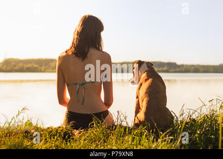 Schöne schlanke Frau mit Hund genießen Sie einen wunderschönen Blick in der Nähe des Sees bei Sonnenuntergang von warmen und sonnigen Nachmittag. Junge Passform weiblichen mit Staffordshire te Sitzung Stockfoto