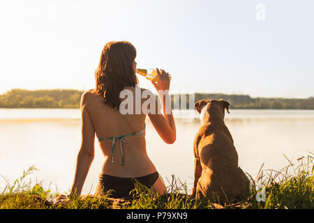 Schöne schlanke Frau mit Hund genießen Sie einen wunderschönen Blick in der Nähe des Sees bei Sonnenuntergang von warmen, sonnigen Nachmittag und einem Drink. Junge Passform weiblichen Sitzen wi Stockfoto