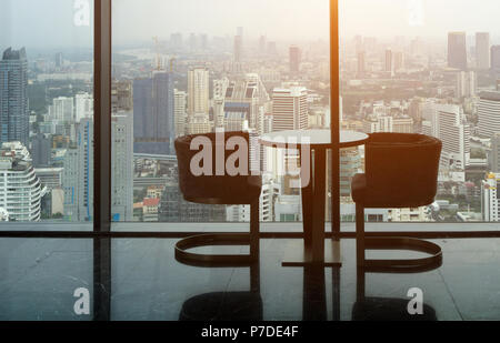 Zwei moderne Sessel und Kreis Glastisch auf Turm Büro mit städtischen Skyline im Hintergrund und die warme Sonne am Nachmittag Zeit. Stockfoto