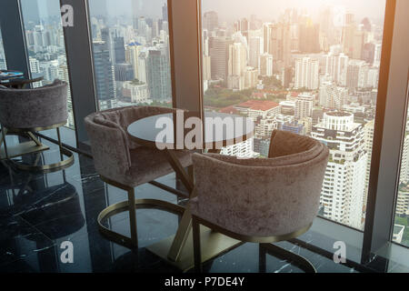 Zwei moderne Sessel und Kreis Glastisch auf Turm Büro mit städtischen Skyline im Hintergrund und die warme Sonne am Nachmittag Zeit. Stockfoto