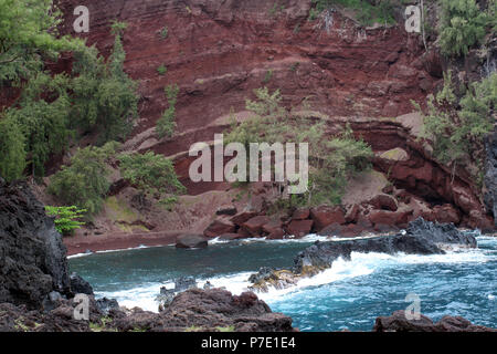 Hoch aufragenden, steilen, roten Felswände führt zu einer Bucht der Red Sand Beach auf Kaihalulu Bay, mit Wellen aus dem Pazifischen Ozean Absturz in Lava Rock i Stockfoto