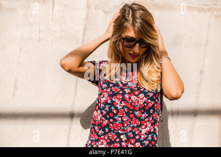 Blond, stilvolle Mitte der erwachsenen Frau Sonnenbrille tragen vor der Wand mit den Händen in das Haar Stockfoto