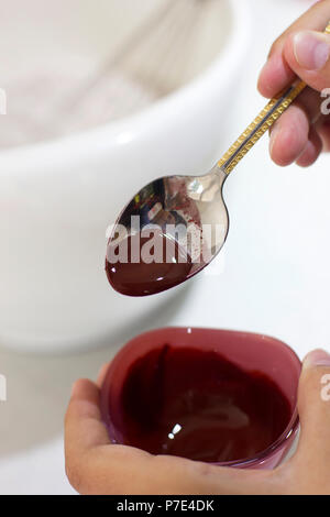 Hände halten einen Löffel und eine Schüssel mit roter Lebensmittelfarbe, die tropft von der Löffel. Stockfoto